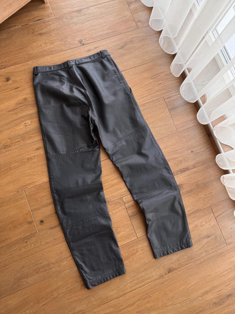 Pu&ograve; includere: Pantaloni in pelle nera dal design classico. I pantaloni hanno un taglio dritto e un'estetica semplice e pulita. La pelle sembra morbida e ha una leggera lucentezza. I pantaloni sono appoggiati su una superficie di legno.