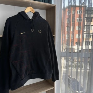 Op de afbeelding: Zwarte hoodie met een rood en wit geometrisch patroon, een Nike swoosh-logo en de tekst "10R" in goud.