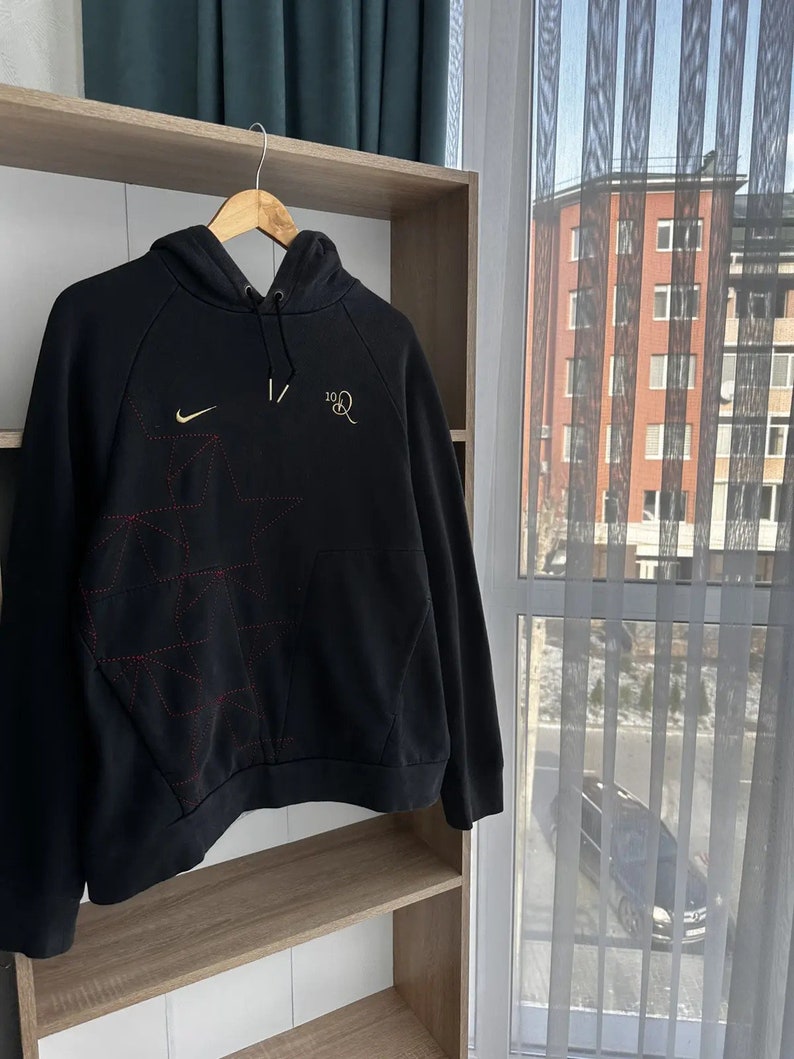 Op de afbeelding: Zwarte hoodie met een rood en wit geometrisch patroon, een Nike swoosh-logo en de tekst "10R" in goud.