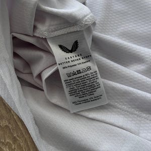 May include: Close-up of a white athletic shirt with a textured pattern. The shirt's tag displays the Castore logo and the text "BETTER NEVER STOPS." The tag also includes washing instructions and fabric composition: 88% Polyester, 12% Elastane.