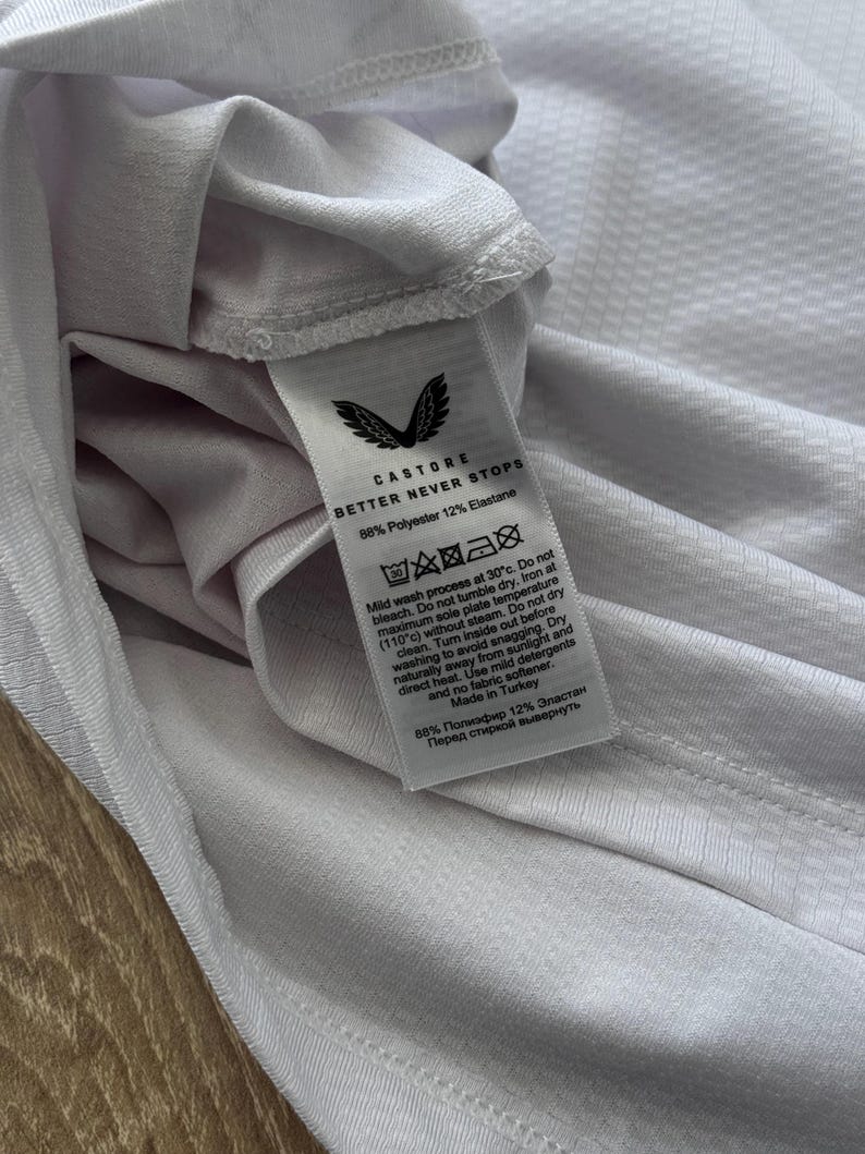 May include: Close-up of a white athletic shirt with a textured pattern. The shirt's tag displays the Castore logo and the text "BETTER NEVER STOPS." The tag also includes washing instructions and fabric composition: 88% Polyester, 12% Elastane.