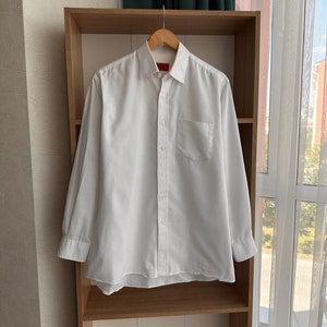 May include: A white long-sleeved button-down shirt with a pocket on the left side. The shirt is hanging on a wooden hanger on a shelf.