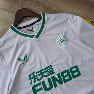 May include: White soccer jersey with green accents, featuring the Newcastle United crest and sponsor logos. The jersey has a V-neck collar with green trim and the Castore logo. The Fun88 logo is prominently displayed in green.