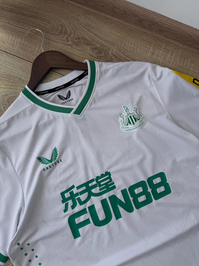 May include: White soccer jersey with green accents, featuring the Newcastle United crest and sponsor logos. The jersey has a V-neck collar with green trim and the Castore logo. The Fun88 logo is prominently displayed in green.