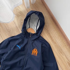 May include: Navy blue hooded jacket with a full zip. The jacket displays a blue Puma logo and a team emblem with the text "DROIT AU BUT" in orange. It has a white interior lining. The jacket is designed for protection from the elements.