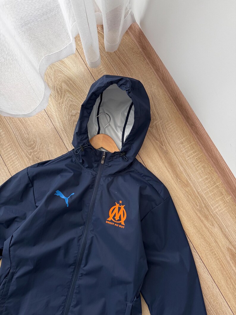 May include: Navy blue hooded jacket with a full zip. The jacket displays a blue Puma logo and a team emblem with the text "DROIT AU BUT" in orange. It has a white interior lining. The jacket is designed for protection from the elements.