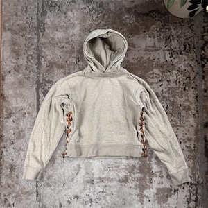 K&ouml;nnte beinhalten: Ein hellgrauer Kapuzenpullover mit Kordelzugkapuze und langen &Auml;rmeln. Die Seiten des Hoodies weisen ein dekoratives Schn&uuml;rdesign mit braunen und orangefarbenen Akzenten auf. Der Hoodie ist an der Taille verk&uuml;rzt.