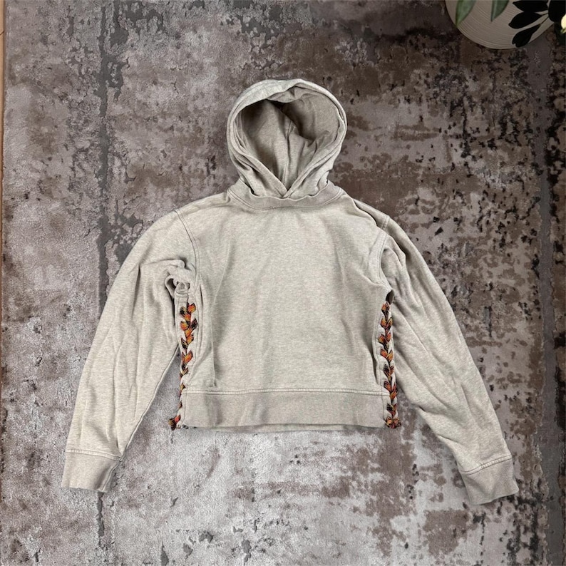K&ouml;nnte beinhalten: Ein hellgrauer Kapuzenpullover mit Kordelzugkapuze und langen &Auml;rmeln. Die Seiten des Hoodies weisen ein dekoratives Schn&uuml;rdesign mit braunen und orangefarbenen Akzenten auf. Der Hoodie ist an der Taille verk&uuml;rzt.