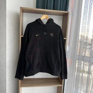Op de afbeelding: Zwarte hoodie met een rood en wit geometrisch ontwerp op de voorkant. De hoodie heeft een klein Nike swoosh-logo op de linkerborst en de tekst "10R" op de rechterborst.
