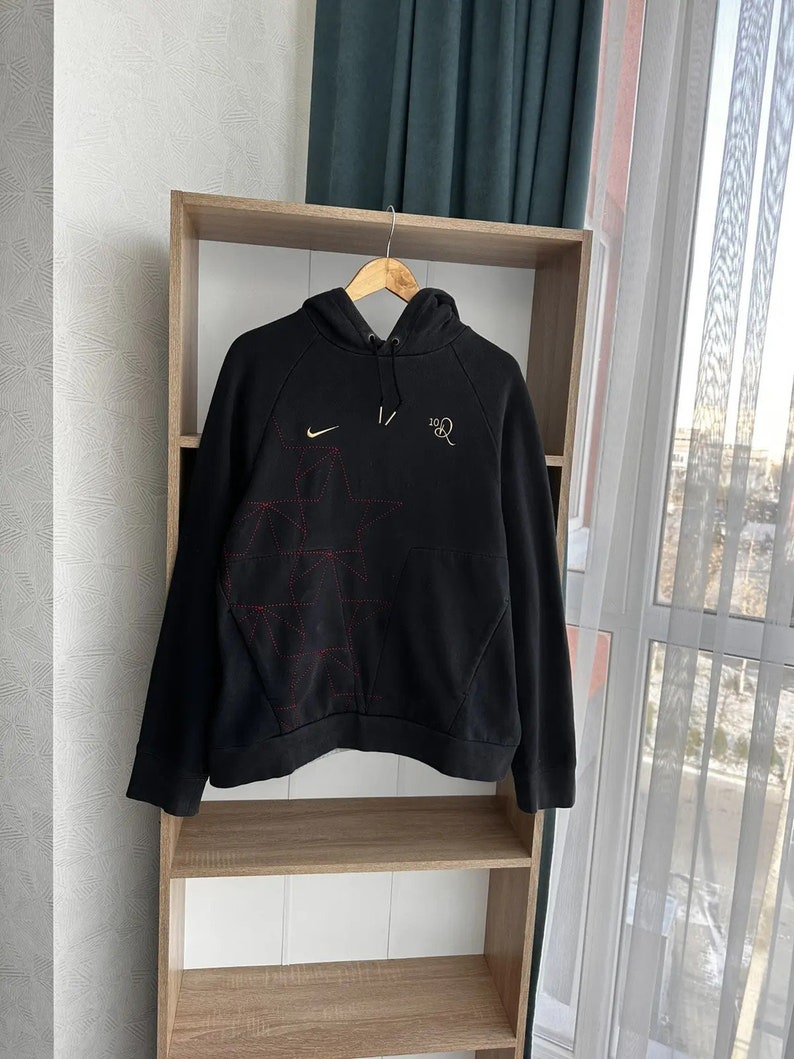 Op de afbeelding: Zwarte hoodie met een rood en wit geometrisch ontwerp op de voorkant. De hoodie heeft een klein Nike swoosh-logo op de linkerborst en de tekst "10R" op de rechterborst.