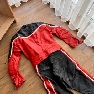 May include: A red and black leather motorcycle racing suit with white stripes. The suit is laid out flat on a wooden floor.