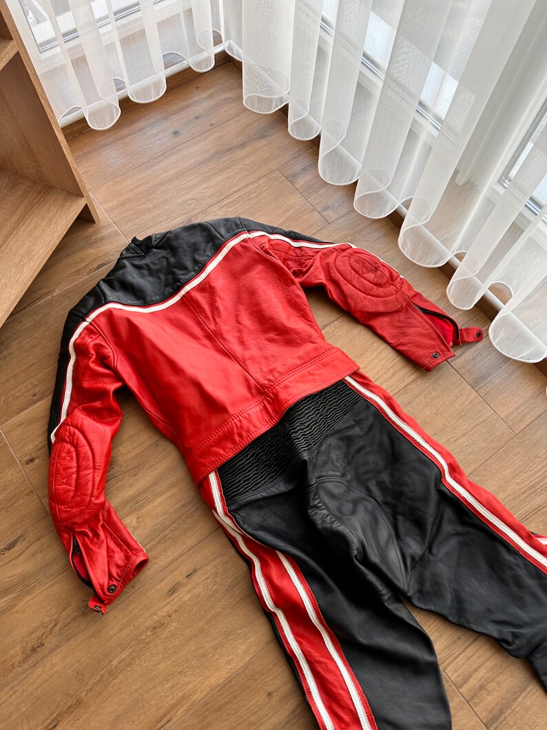 May include: A red and black leather motorcycle racing suit with white stripes. The suit is laid out flat on a wooden floor.