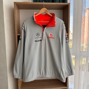 Può includere: Una giacca in pile grigia con una cerniera rossa e dettagli arancioni. La giacca ha il logo Mercedes-Benz, il logo Mobil 1 e il logo Vodafone sul davanti.