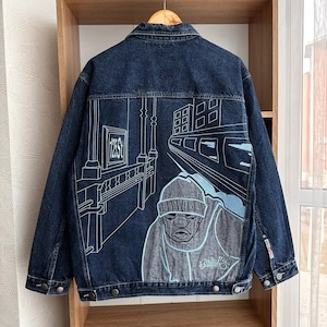 Könnte beinhalten: Dunkelblaue Jeansjacke mit Grafikdesign auf dem Rücken. Das Design zeigt eine U-Bahnszene mit einer Figur mit Mütze und dem Text "125 St". Die Jacke hat einen klassischen Kragen, einen Knopfverschluss und Taschen.