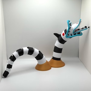 Pode incluir: Um brinquedo segmentado preto e branco com uma cabeça azul e branca em forma de tubarão. O brinquedo está posicionado com a cabeça voltada para o lado direito da imagem. O brinquedo está sobre uma superfície branca.