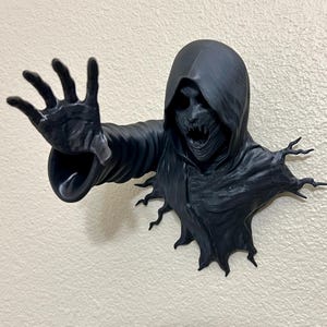 以下が含まれることがあります： フードをかぶった人物の壁掛け彫刻。黒い3Dプリントで、手を伸ばし、脅迫的な表情をしている。