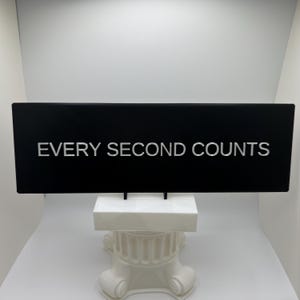 以下が含まれることがあります： 「EVERY SECOND COUNTS」の白い文字が書かれた黒い長方形の看板。白い装飾的な台座の上に設置され、白い背景に配置されています。