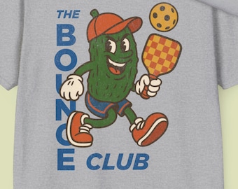May include: A grey t-shirt featuring a cartoon pickle playing pickleball. The pickle is wearing a blue and white baseball cap and holding a paddle. The text "The Bounce Club" is printed on the shirt.
