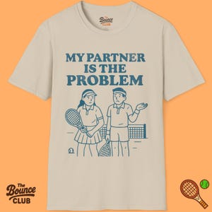 May include: A cream-coloured t-shirt with the blue text "MY PARTNER IS THE PROBLEM". The design includes a cartoon of two tennis players, a net, and a tennis ball. The Bounce Club logo is displayed at the top and bottom.
