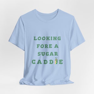 May include: Light blue t-shirt with the text "LOOKING FORE A SUGAR CADDIE" in green letters. A cartoon golf bag with a golf club is next to the text.