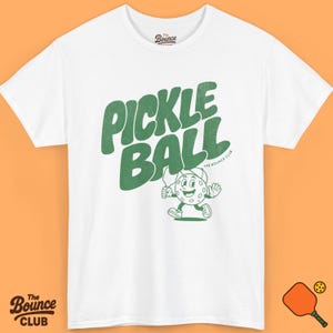 Puede incluir: Camiseta blanca con la inscripción "Pickle Ball" en verde y un personaje de pepinillo de dibujos animados sosteniendo una raqueta. El logotipo de The Bounce Club está impreso arriba y abajo.