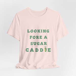 May include: A pink t-shirt with the text "LOOKING FORE A SUGAR CADDIE" in green letters. A green and yellow golf bag icon is next to the text.