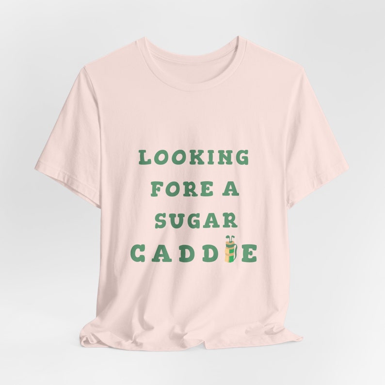 May include: A pink t-shirt with the text "LOOKING FORE A SUGAR CADDIE" in green letters. A green and yellow golf bag icon is next to the text.