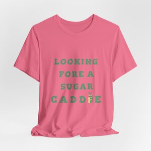 May include: Pink t-shirt with the text "LOOKING FORE A SUGAR CADDIE" in green text. A green and yellow golf bag with a golf club is next to the word "CADDIE".