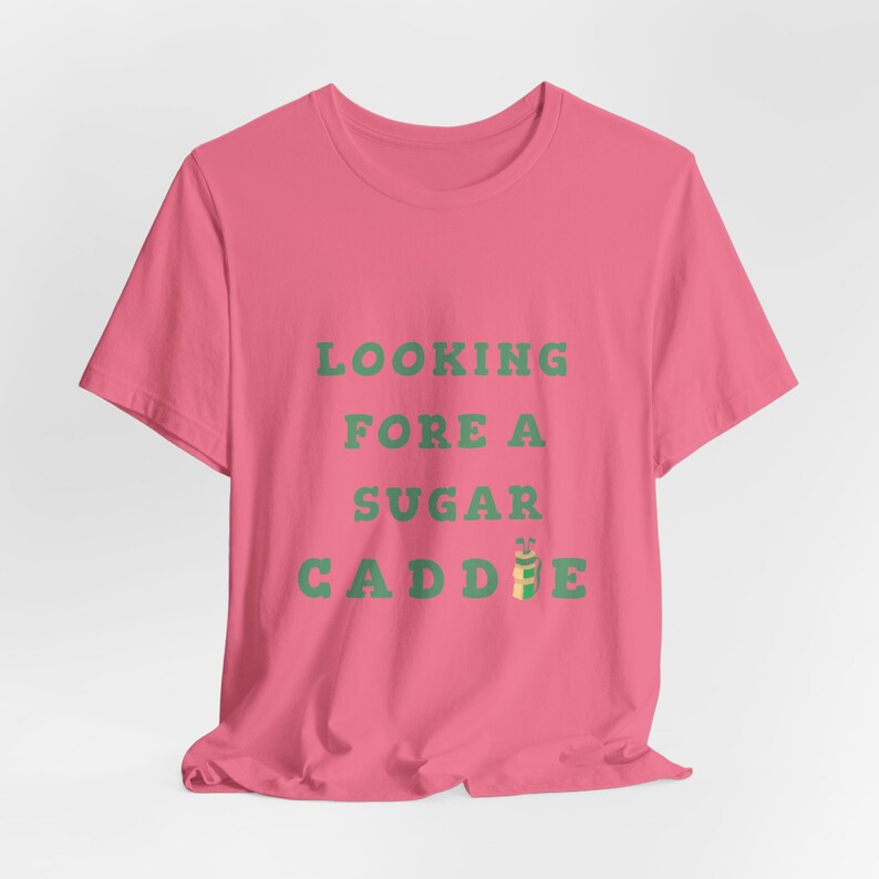 May include: Pink t-shirt with the text "LOOKING FORE A SUGAR CADDIE" in green text. A green and yellow golf bag with a golf club is next to the word "CADDIE".