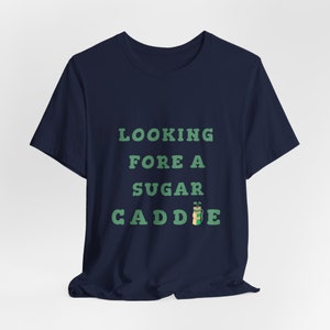 May include: A navy blue t-shirt with the text "LOOKING FORE A SUGAR CADDIE" in green text with a cartoon golf bag and clubs.
