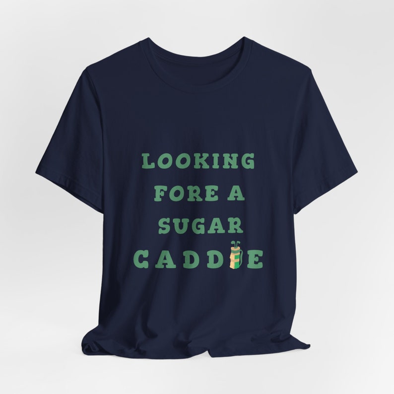 May include: A navy blue t-shirt with the text "LOOKING FORE A SUGAR CADDIE" in green text with a cartoon golf bag and clubs.