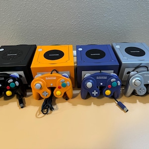 Könnte beinhalten: Vier Nintendo GameCube Konsolen und passende Controller in Schwarz, Orange, Blau und Silber. Jede Konsole hat ein kreisförmiges Laufwerk und einen Power-Knopf. Die Controller haben ein klassisches Tastenlayout.
