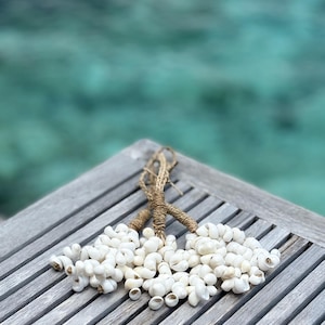 May include: Two strands of white seashells are tied together with natural rope. The seashells are clustered together in a decorative arrangement. The background is a blurred view of turquoise water.
