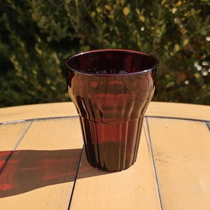 May include: A dark red glass tumbler with vertical ribbed detailing. The glass is empty and sits on a light brown wooden table. The background features green foliage, and the lighting suggests a sunny day.