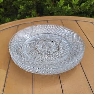May include: A stack of clear, cut-glass plates and a smaller serving dish. The plates have a textured, diamond-patterned rim and a smooth center. The serving dish has a starburst design in the center. The plates are stacked on a wooden table.