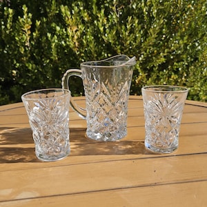 May include: A clear glass pitcher with a handle and two matching glasses. The pitcher and glasses have a diamond and starburst pattern. The set is displayed on a wooden table with a green leafy background.