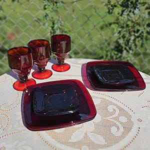 May include: A set of vintage ruby red glassware, including three stemmed glasses and two square plates. The glasses and plates are arranged on a white lace tablecloth, with a blurred green background.