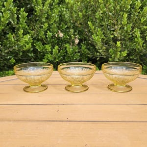 May include: Three yellow glass dessert bowls with a floral pattern. The bowls have a rounded shape and sit on a short pedestal base. The bowls are arranged in a row on a wooden surface, with a green leafy background.