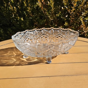 May include: A clear, oval-shaped glass bowl with a decorative, textured pattern and three small feet. The bowl has a scalloped rim and is sitting on a wooden surface. The bowl is likely used for serving or display.