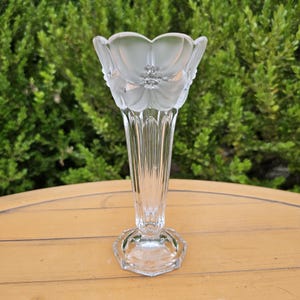 May include: A clear glass vase featuring a flared top rim with a frosted floral pattern. The body of the vase has vertical fluted ridges that taper down to a hexagonal base. The glass is transparent with a smooth texture on the lower section.