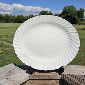 May include: A white oval serving platter with a swirled design around the rim. The platter is displayed on a black stand, set against a backdrop of green grass, trees, and a blue sky with clouds.