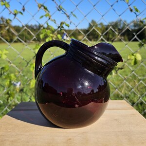 May include: A dark purple glass pitcher with a rounded body, a handle, and a pouring spout. The pitcher is sitting on a wooden surface, with a chain-link fence and greenery in the background. The pitcher is approximately 10 inches tall.
