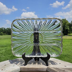May include: A clear, rectangular glass serving dish with three compartments, resting on a black metal stand. The dish features a sunburst design and a textured, ridged border. The background shows a green lawn and a blue sky with clouds.