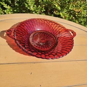 May include: A ruby red glass serving dish with a textured, honeycomb-like pattern. The dish has a circular shape with a central design and two handles. The dish is likely vintage and suitable for serving food.