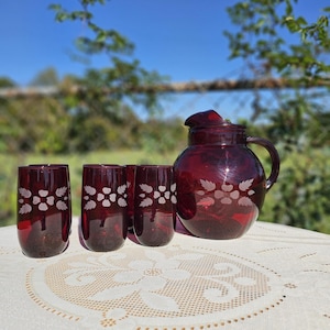 May include: A ruby red glass pitcher and matching tumblers. The pitcher has a handle and a lid. The glasses and pitcher are decorated with a white floral design. The set is displayed on a white lace tablecloth.