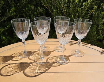 May include: Six clear crystal wine glasses with a textured, etched design. The glasses have a stemmed base and a round foot. The glasses are arranged on a light brown wooden surface, with a green leafy background.