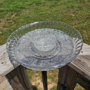 May include: A clear glass serving set with a large, round, fluted-edge platter and a smaller bowl nested inside. The platter and bowl have embossed floral and geometric patterns. The set is sitting on a wooden surface.