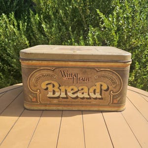 May include: A rectangular metal bread box with a tan and brown finish. The front features the text Wheat Heart Brand Bread, Oven-Baked, Enriched, and The Finest Quality Since 1845, decorated with wheat stalk illustrations and ornate borders.