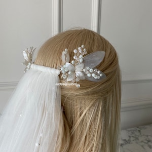Op de afbeelding: Een witte bruidskapselkam met parel- en kristallen versieringen. De kam is versierd met delicate witte bloemen en bladeren. De kam is te zien in het haar van een model met een witte sluier.