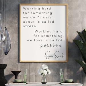 Könnte beinhalten: Ein gerahmtes Zitat von Simon Sinek, das lautet: "Hart zu arbeiten für etwas, das uns nicht interessiert, nennt man Stress. Hart zu arbeiten für etwas, das wir lieben, nennt man Leidenschaft." Das Zitat ist auf weißem Hintergrund mit einem Holzrahmen dargestellt. Der Rahmen hängt an einer grauen Wand über einem Tisch mit einer Vase mit weißen Blumen.